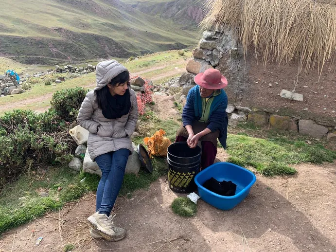 Two women outdoors in a rural, mountainous area. One woman in a gray hooded jacket and jeans sits on rocks; the other woman, wearing a pink hat, is seated next to a bucket and blue tub.