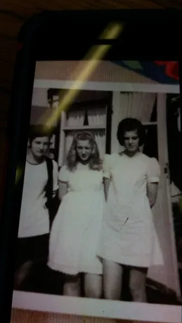 A somewhat blurry, black and white scanned photograph featuring three individuals. Two figures are in dresses, and one is wearing a light-colored shirt, possibly a t-shirt.