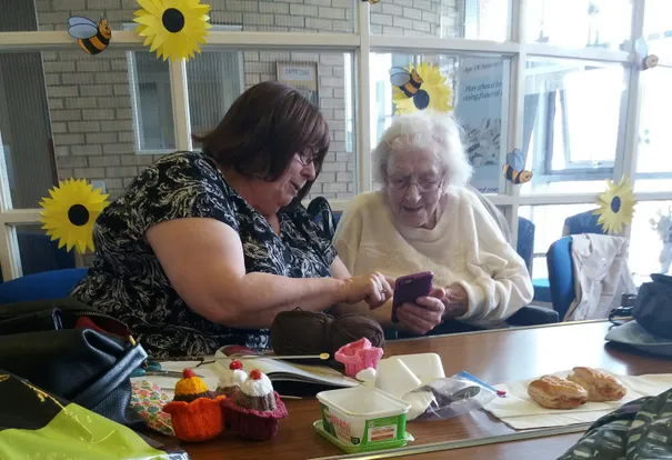 A photograph shows two women sitting at a table, with one woman in a black patterned top looking at the other, who is holding a phone. The table is covered with yarn, knitted crafts, packaged foods, and pastries.